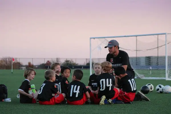 Youth sports boys soccer team sitting in a huddle with their coach teaching life lessons and sports skills on a soccer field